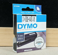 Dymo white plastic label refill tape in its original retail blister packaging, featuring a black tape roll inside a clear plastic casing on a green and white card backing.