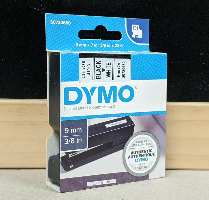 Dymo white plastic label refill tape in its original retail blister packaging, featuring a black tape roll inside a clear plastic casing on a green and white card backing.