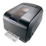 A Honeywell thermal label printer in matte black, shown with a barcode label partially ejected from the front. The printer features a glossy black top surface with the Honeywell logo, a translucent window on the lid, and a red power button.