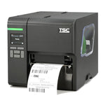 A TSC industrial label printer in matte black, shown from a three-quarter angle with a white label sheet partially ejected from the front slot. The printer features a small display screen on the left, a green status light, and a clear side window.