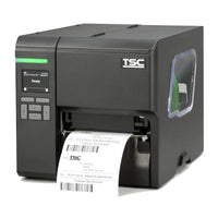 A TSC industrial label printer in matte black, shown from a three-quarter angle with a white label sheet partially ejected from the front slot. The printer features a small display screen on the left, a green status light, and a clear side window.