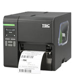 A TSC industrial barcode label printer with a dark gray and silver metallic body, showing a digital control screen on the front left and a white label sheet being printed and ejected from the front right.