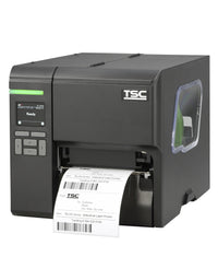 A TSC industrial barcode label printer with a dark gray and silver metallic body, showing a digital control screen on the front left and a white label sheet being printed and ejected from the front right.