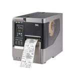 A TSC industrial barcode label printer with a dark gray and silver metallic body, showing a digital control screen on the front left and a white label sheet printing out from the front right.