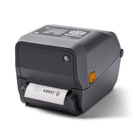 A Zebra thermal label printer in dark gray with a small orange indicator light, shown with a white label partially ejected from the front on a white background.